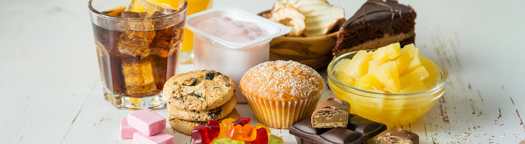 Assortment of sugary foods and drinks including soda, cookies, muffin, yogurt, gummy candies, chocolate, cake, and pineapple pieces on a table.