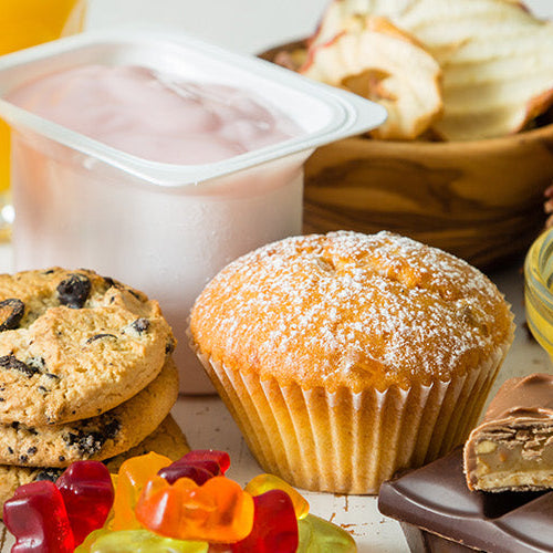 Assortment of sugary foods and drinks including soda, cookies, muffin, yogurt, gummy candies, chocolate, cake, and pineapple pieces on a table.