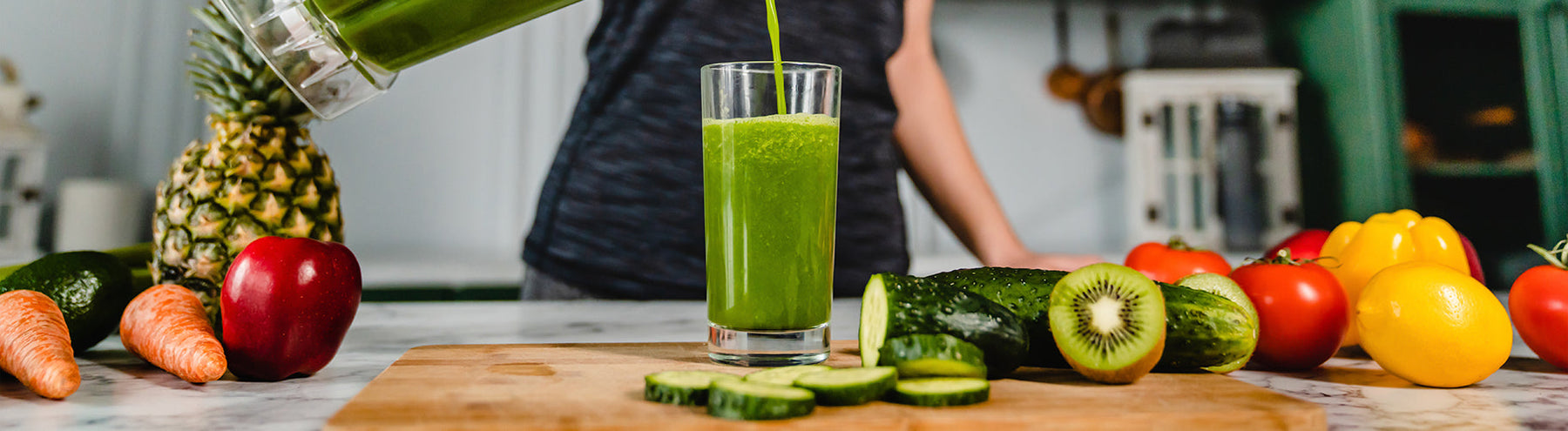 Fresh green smoothie being poured into a glass beside cucumbers, kiwi, pineapple, carrots, tomatoes, and other fresh produce.