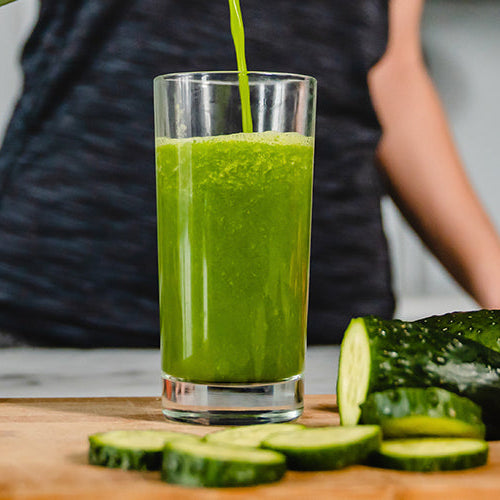 Fresh green smoothie being poured into a glass beside cucumbers, kiwi, pineapple, carrots, tomatoes, and other fresh produce.